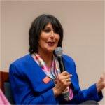 President Mantella speaks into a microphone during a panel discussion. She wears a blue blazer, pink scarf, and a pin that reads �Tech Week�.